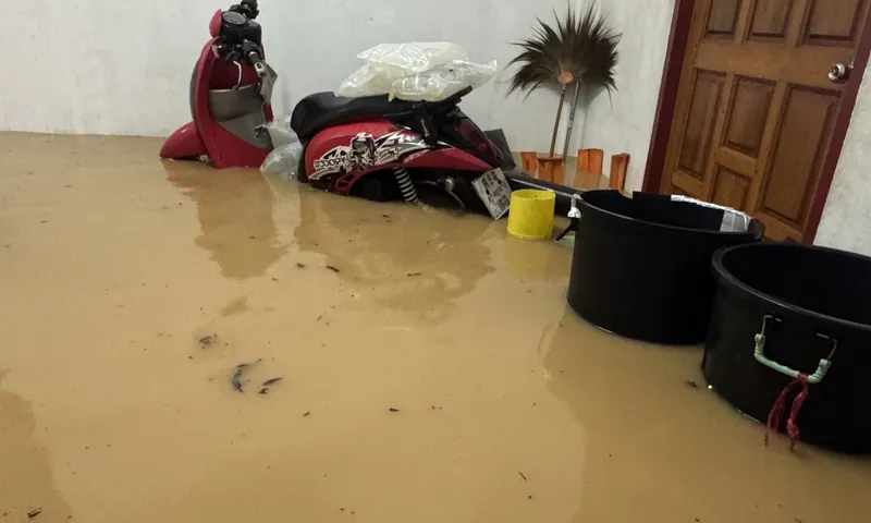 Floodwater inside a home in Thailand — a covered scooter and household containers stand in brown murky water on a ground-floor room.